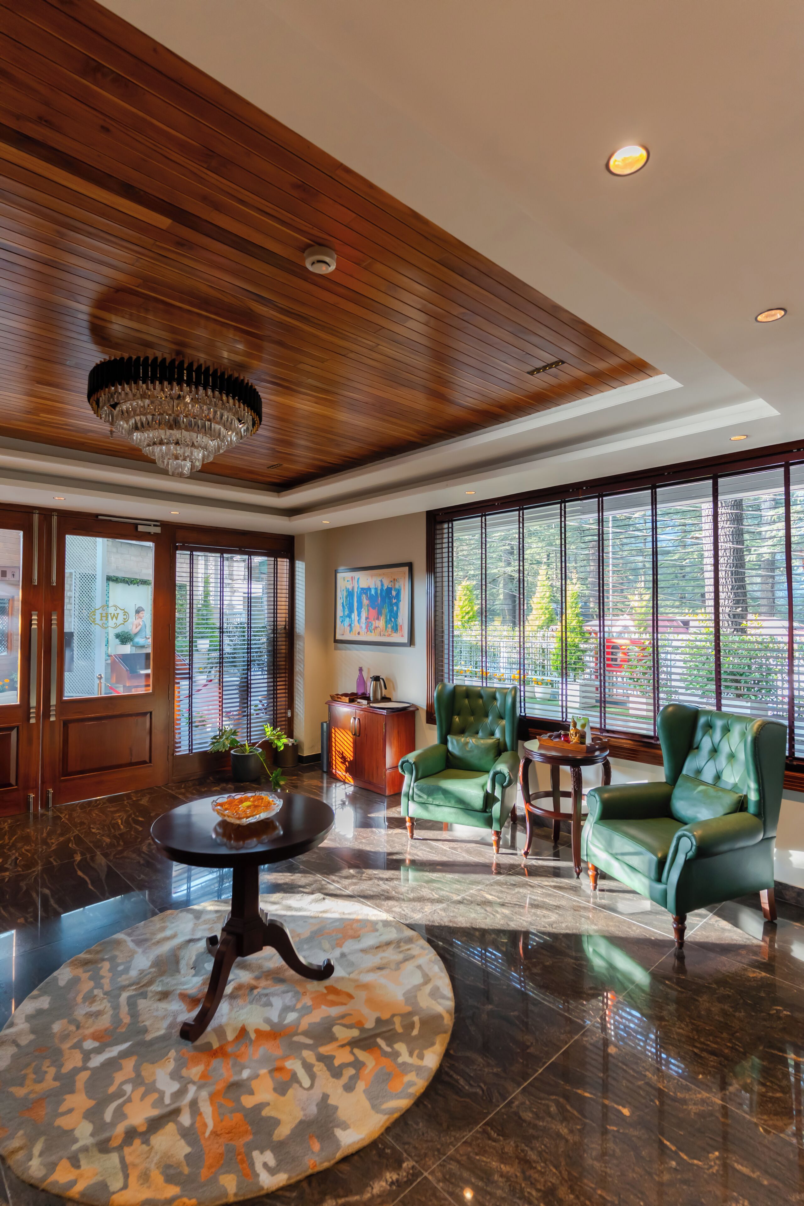 A lobby with a wooden ceiling, large windows, green leather chairs, a round table, and a chandelier.