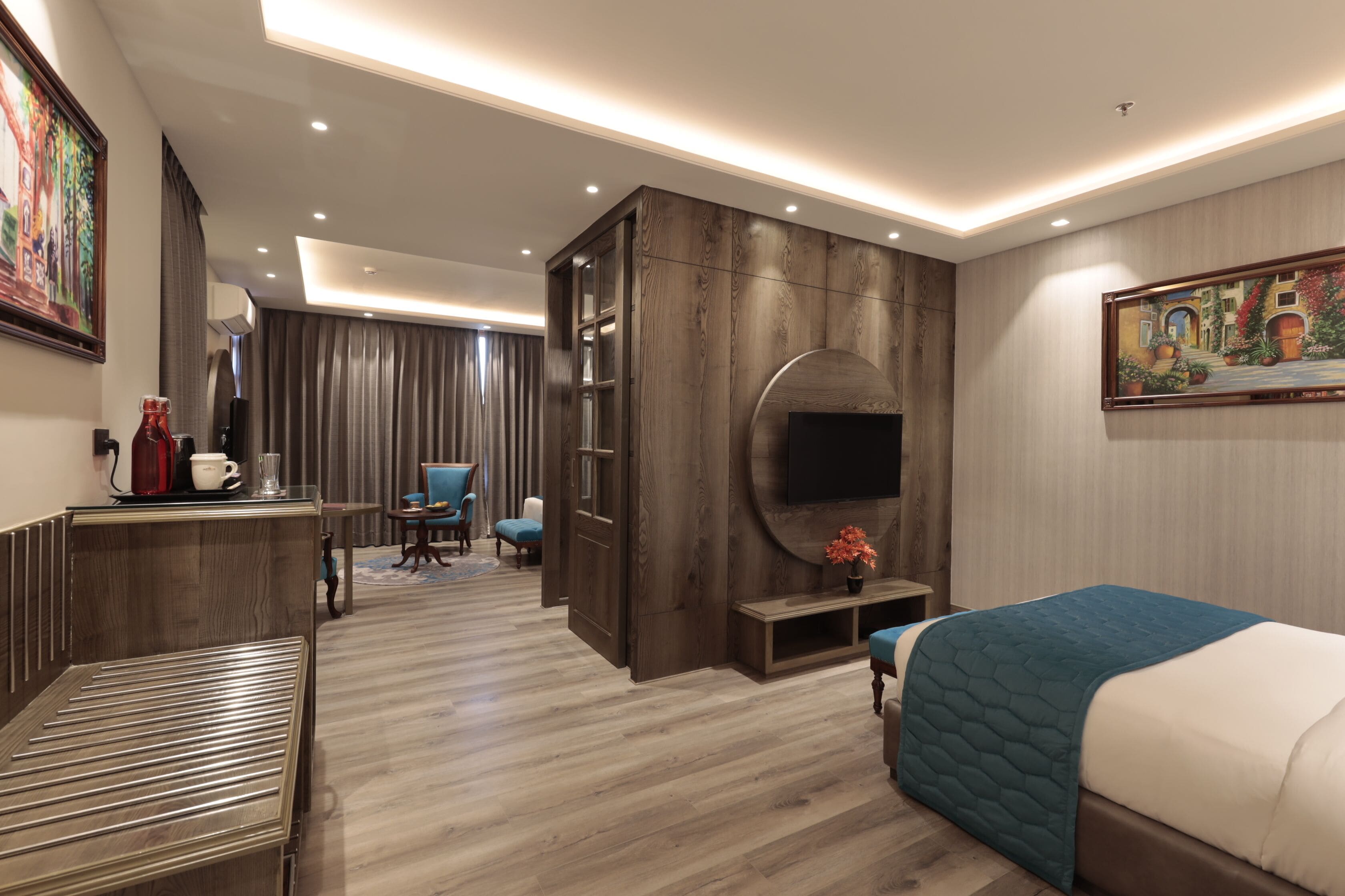 A modern hotel room with a wooden floor, a bed with a blue quilt, a TV mounted on the wall, a desk with a coffee maker, and a chair.