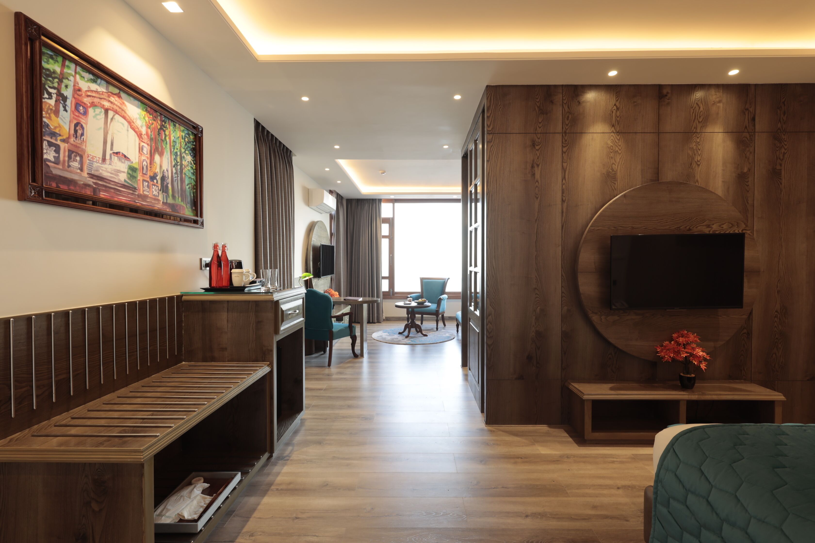 A hotel room with a wooden bench, a desk with a computer, a TV mounted on the wall, and a large window.
