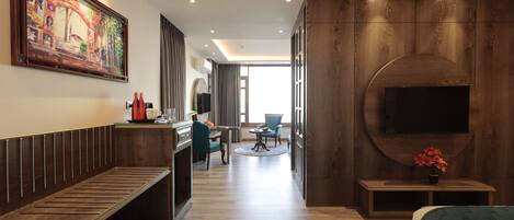 A hotel room with a wooden bench, a desk with a computer, a TV mounted on the wall, and a large window.