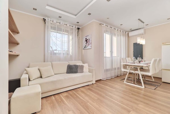 A modern living room with a sofa, dining table, and chairs, wooden flooring, and a ceiling with recessed lighting.