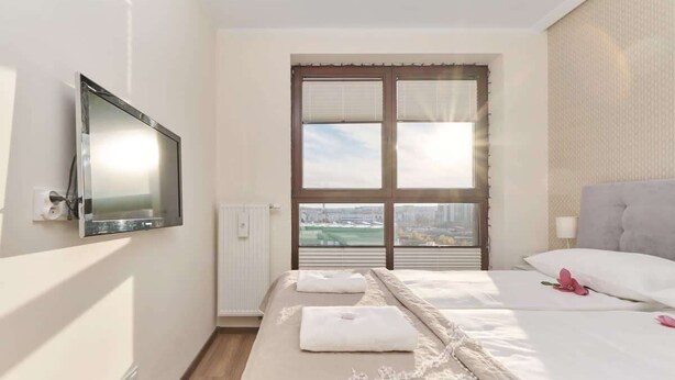 A hotel room with a bed, a TV mounted on the wall, a window with blinds, and a view of the cityscape.