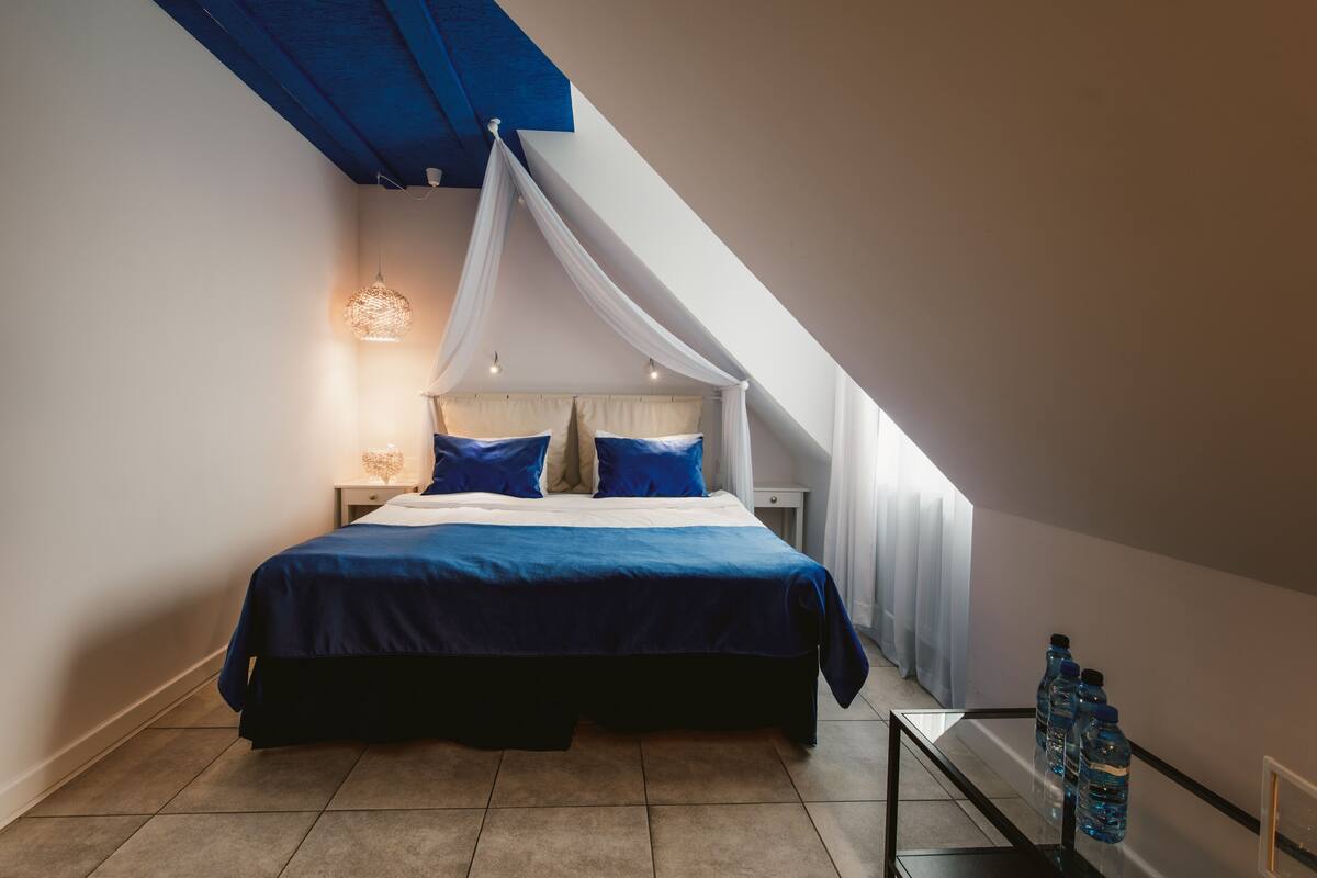 Un dormitorio con cama, mesita de noche y velador. Hay una ventana con cortinas, un techo con una viga azul y una botella de agua sobre una mesa.