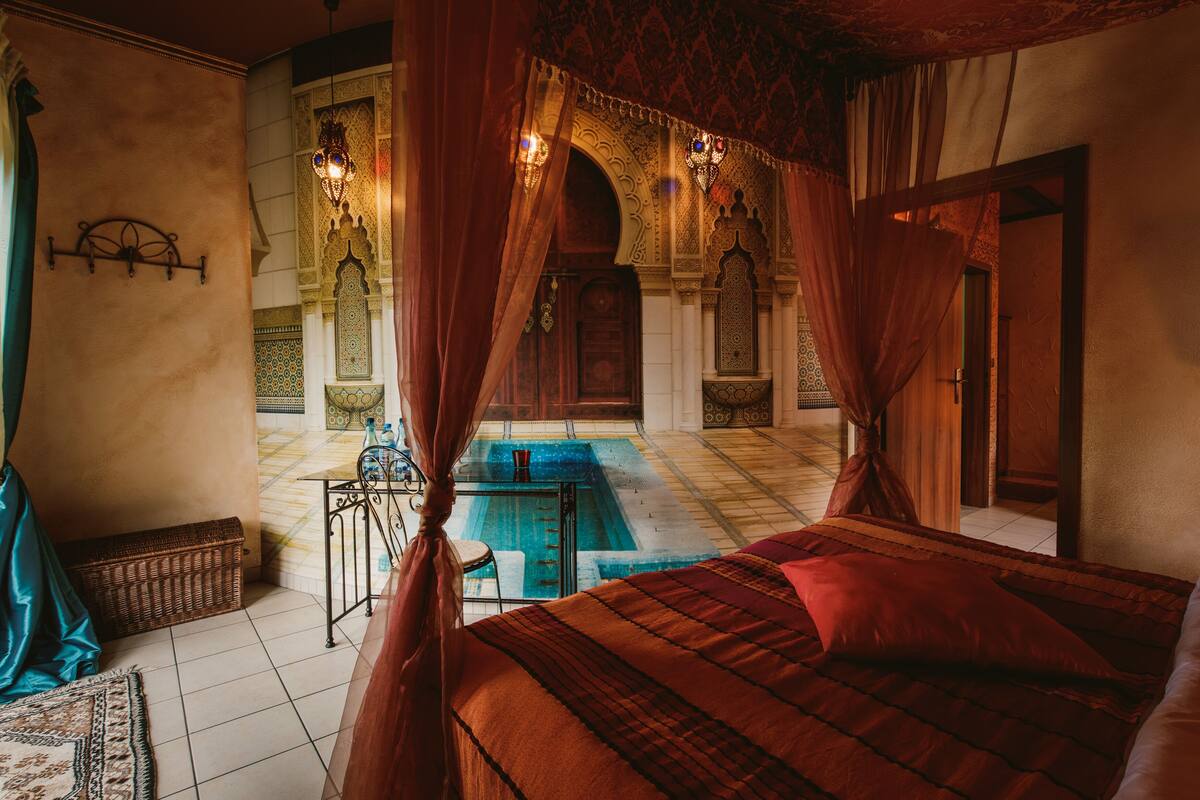 Un dormitorio con una cama grande, una mesita con una silla, una lámpara decorativa en la pared y vistas a un patio exterior con piscina.