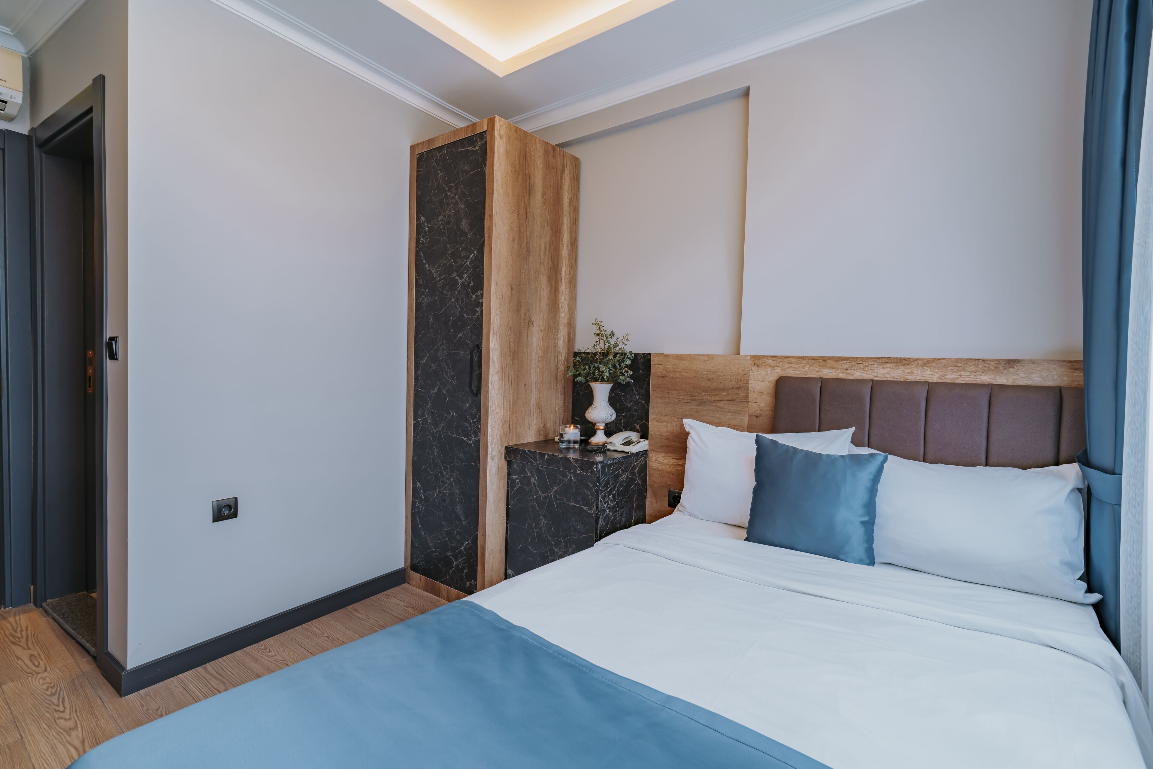 A modern hotel room with a large bed, a wooden headboard, a marble accent wall, and a small bedside table with a lamp.