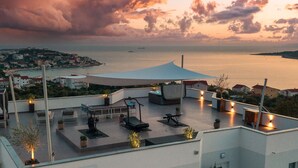 Eine Dachterrasse mit Fitnessgeräten, Sitzbereichen und Blick auf das Meer und die Stadt im Sonnenuntergang.