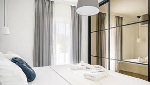 Ein modernes Hotelzimmer mit einem großen Bett, weißen Leinentextilien, Blick durch eine Glastür und einer hängenden Pendelleuchte.