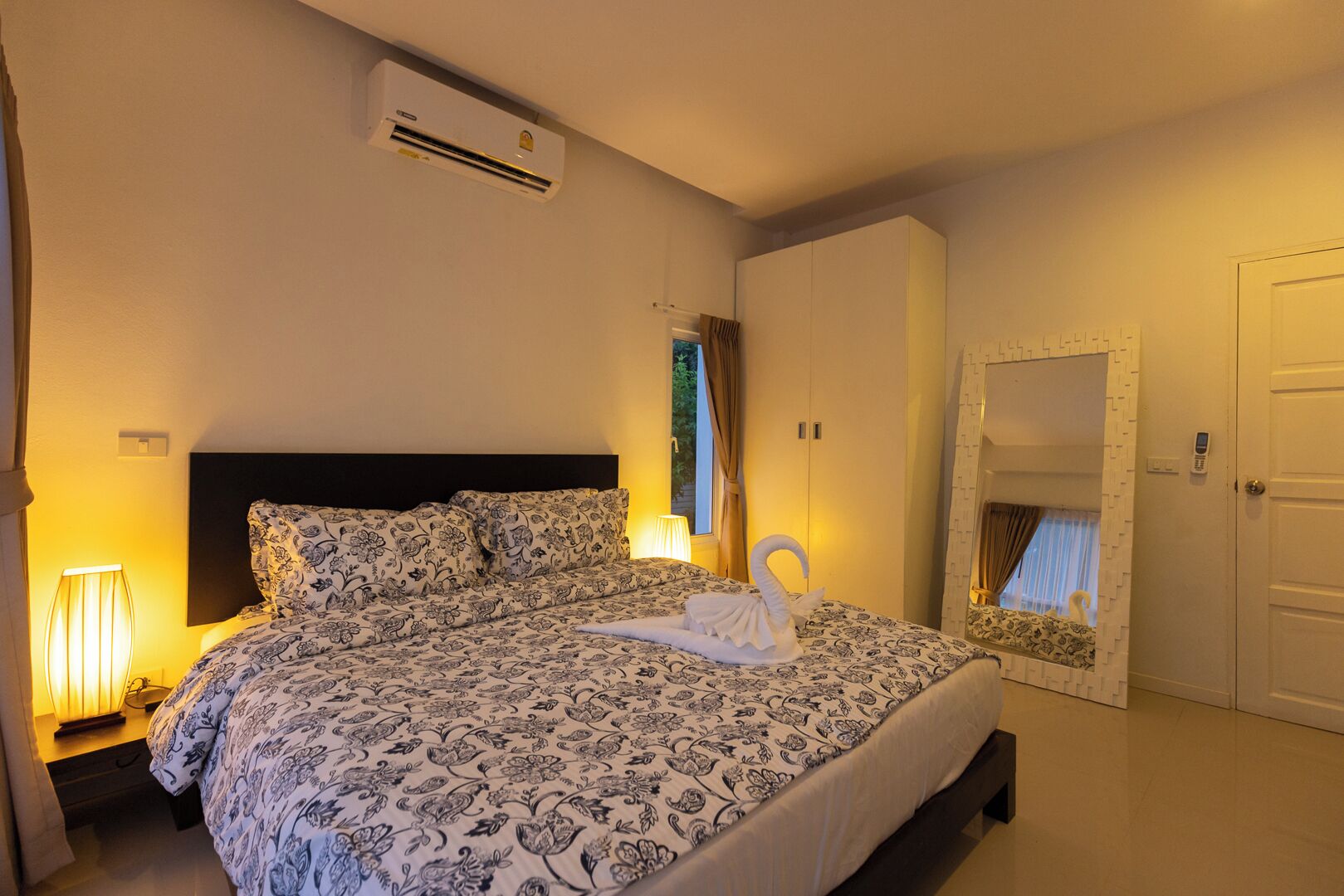 A bedroom with a bed, bedside lamps, an air conditioning unit, a wardrobe, and a mirror.