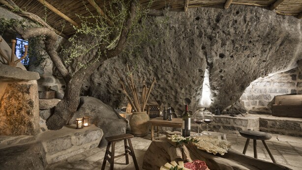 A rustic interior with stone walls, a wooden table with cheese and wine, and a large tree trunk. - Gallicantu Stazzo Retreat (Luogosanto)