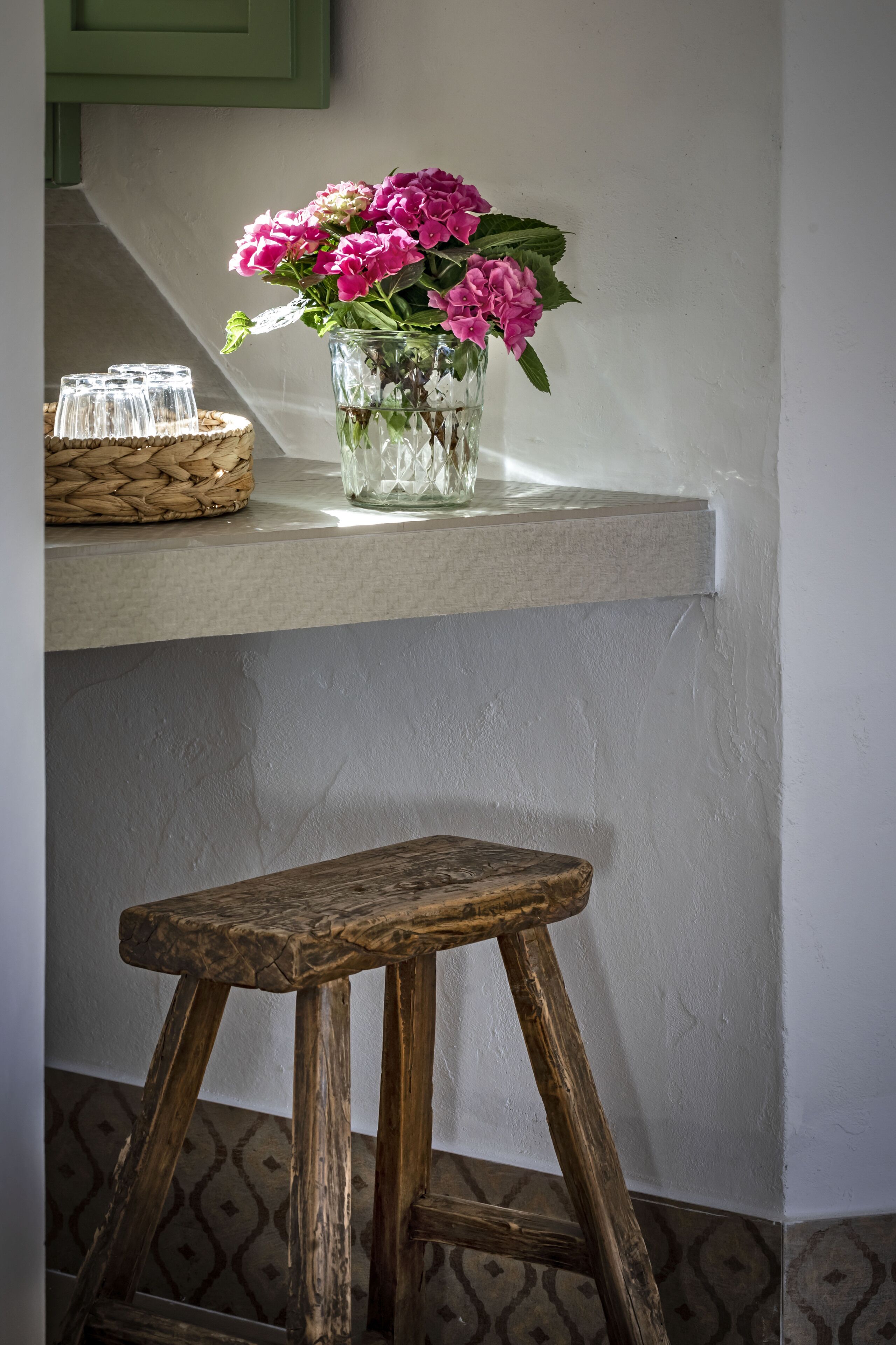 A wooden stool with a woven basket beside a vase of pink flowers on a shelf.