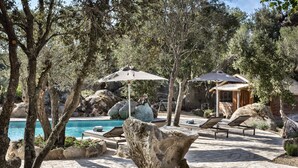 A pool area with lounge chairs and umbrellas, surrounded by rocks and trees. - Gallicantu Stazzo Retreat (Luogosanto)