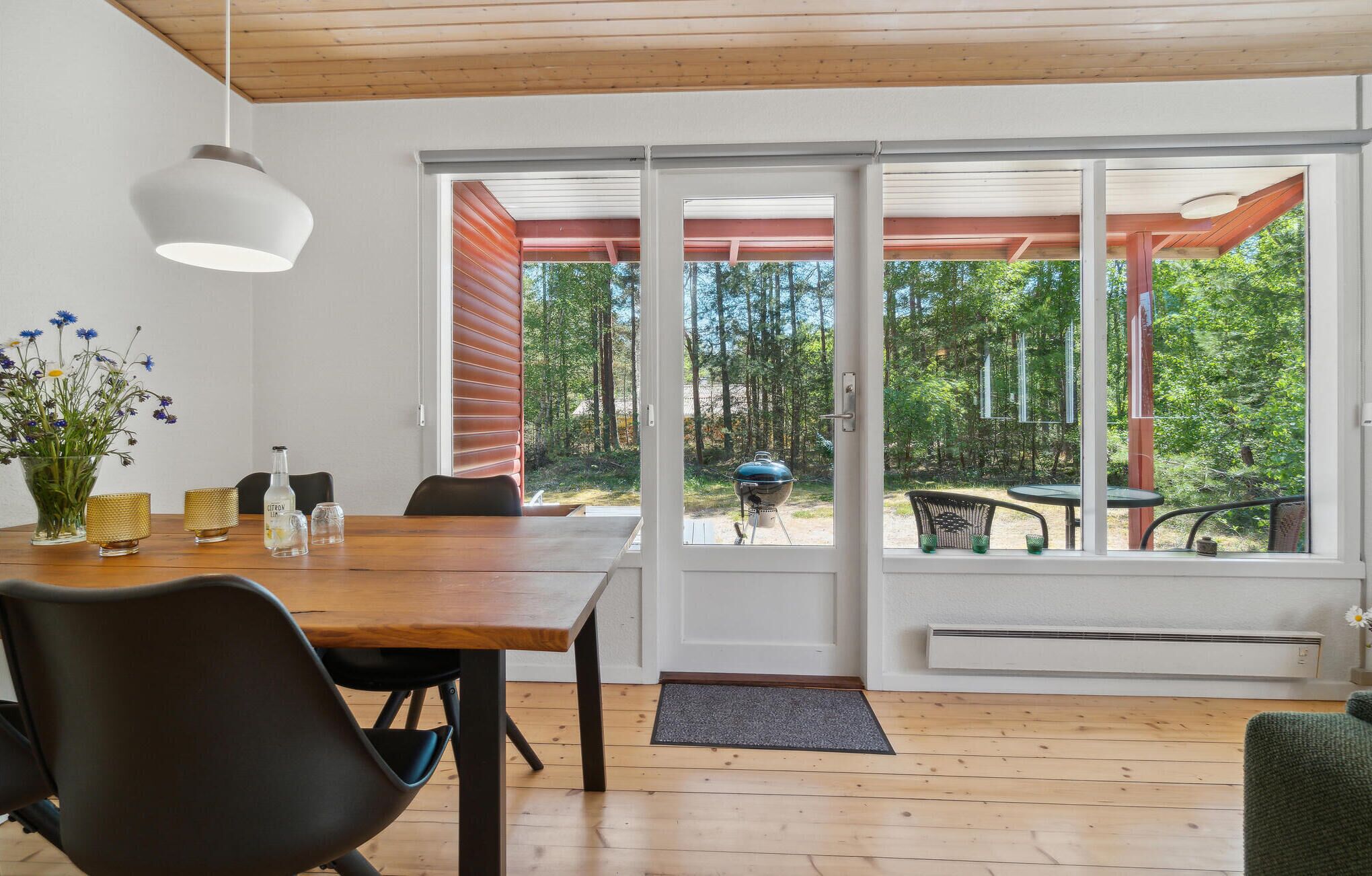 Comedor con mesa de madera, sillas y vista a un bosque a través de amplias puertas correderas de vidrio.