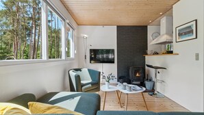 A modern living room with a fireplace, a sofa, a TV, and a view of trees through large windows. - Nice home in Aakirkeby with WiFi (Aakirkeby)