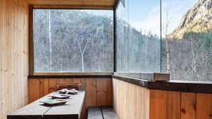 A wooden cabin with a large window overlooking a forested mountain. Inside, there is a wooden table with several dishes and a bench.