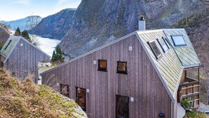 A modern wooden cabin with a sloped roof, multiple windows, and a view of a mountainous landscape with a lake.