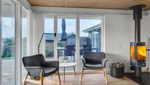 Une pièce moderne dotée d’une grande fenêtre, de deux fauteuils gris rembourrés, d’une table ronde en bois et d’un poêle à bois.