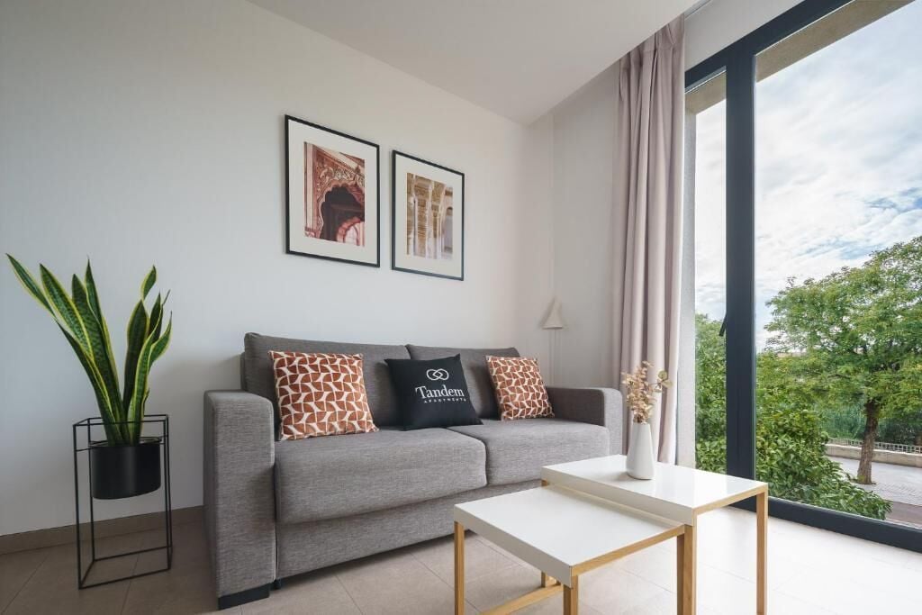 A modern living room with a grey sofa, two framed pictures, a potted plant, and a view of greenery through large windows.