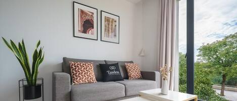 A modern living room with a grey sofa, two framed pictures, a potted plant, and a view of greenery through large windows.
