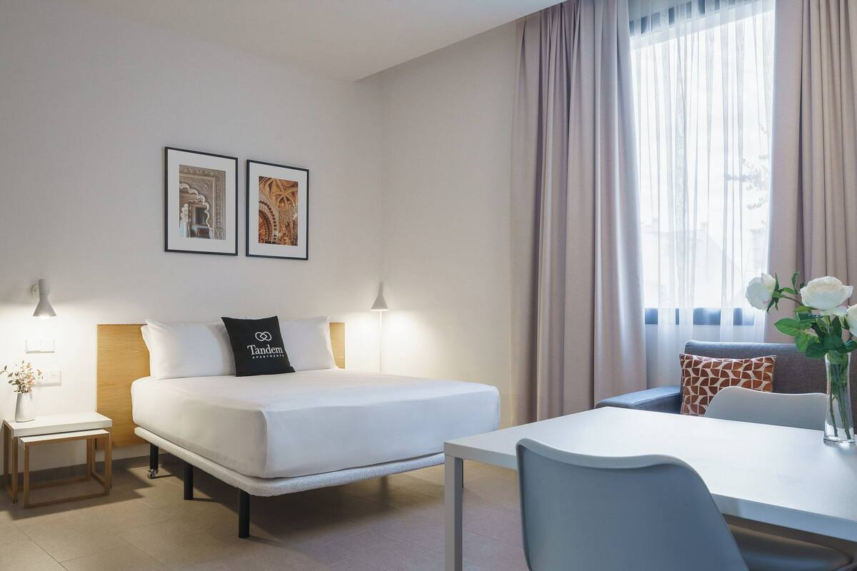 Een moderne hotelkamer met een bed, nachtkastjes, een bureau en een stoel. Er hangen twee ingelijste foto’s aan de muur, een raam met gordijnen en een vaas met bloemen op het bureau.