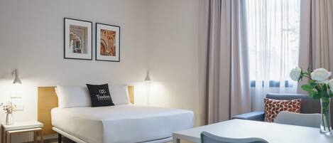 A modern hotel room with a bed, bedside tables, a desk, and a chair. There are two framed pictures on the wall, a window with curtains, and a vase with flowers on the desk.
