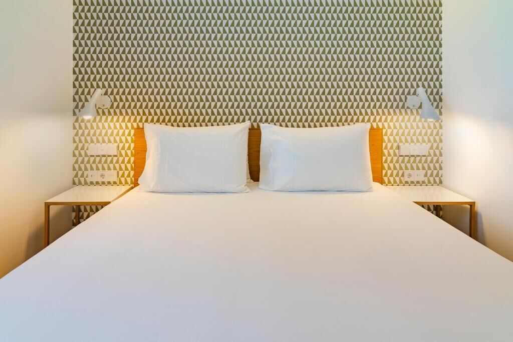 Een bed met een hoofdbord met een geometrisch patroon, twee nachtkastjes met lampen en een effen witte spreidekker.