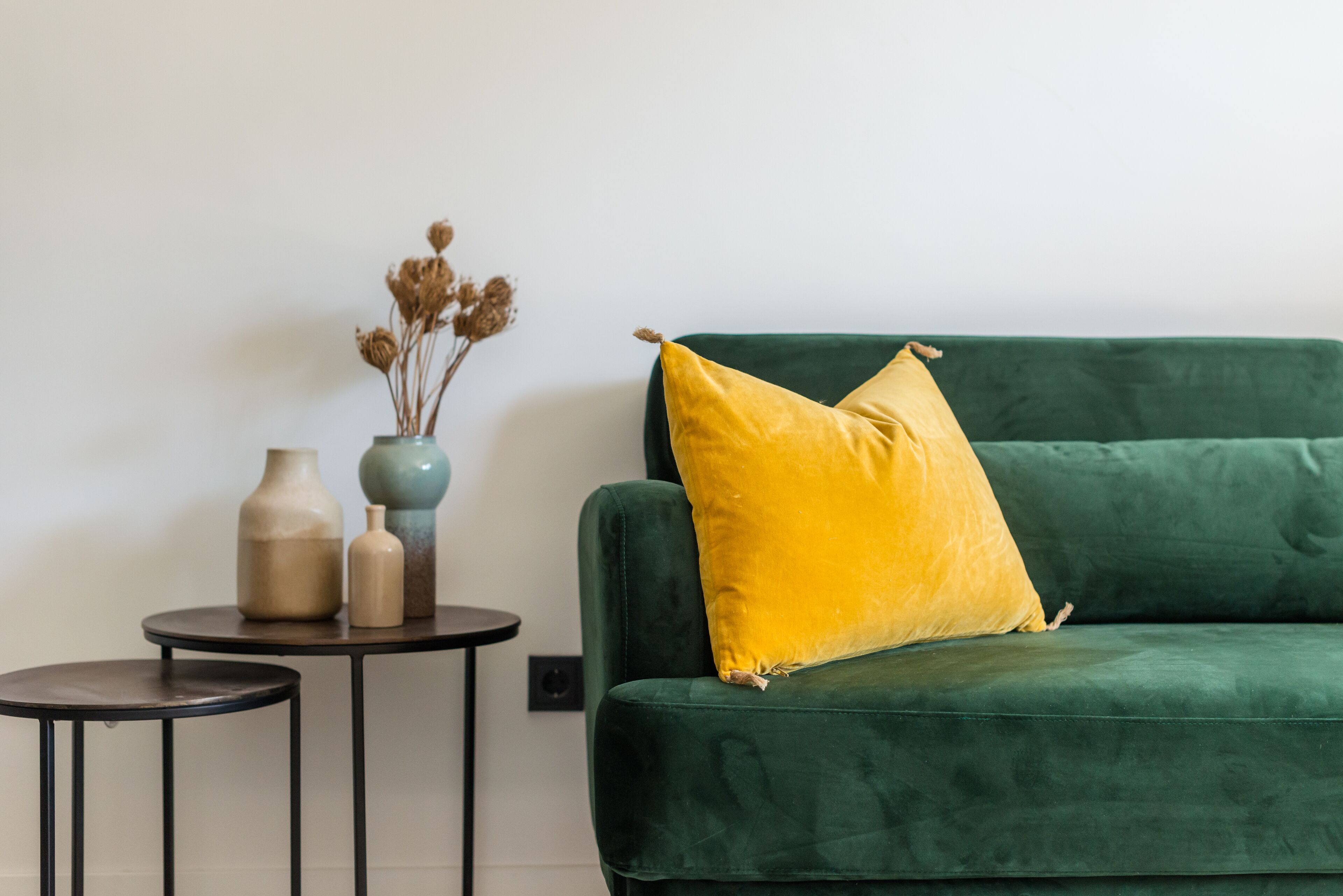 Un sofá verde con un cojín amarillo, una mesita redonda con dos jarrones y flores secas en la pared.