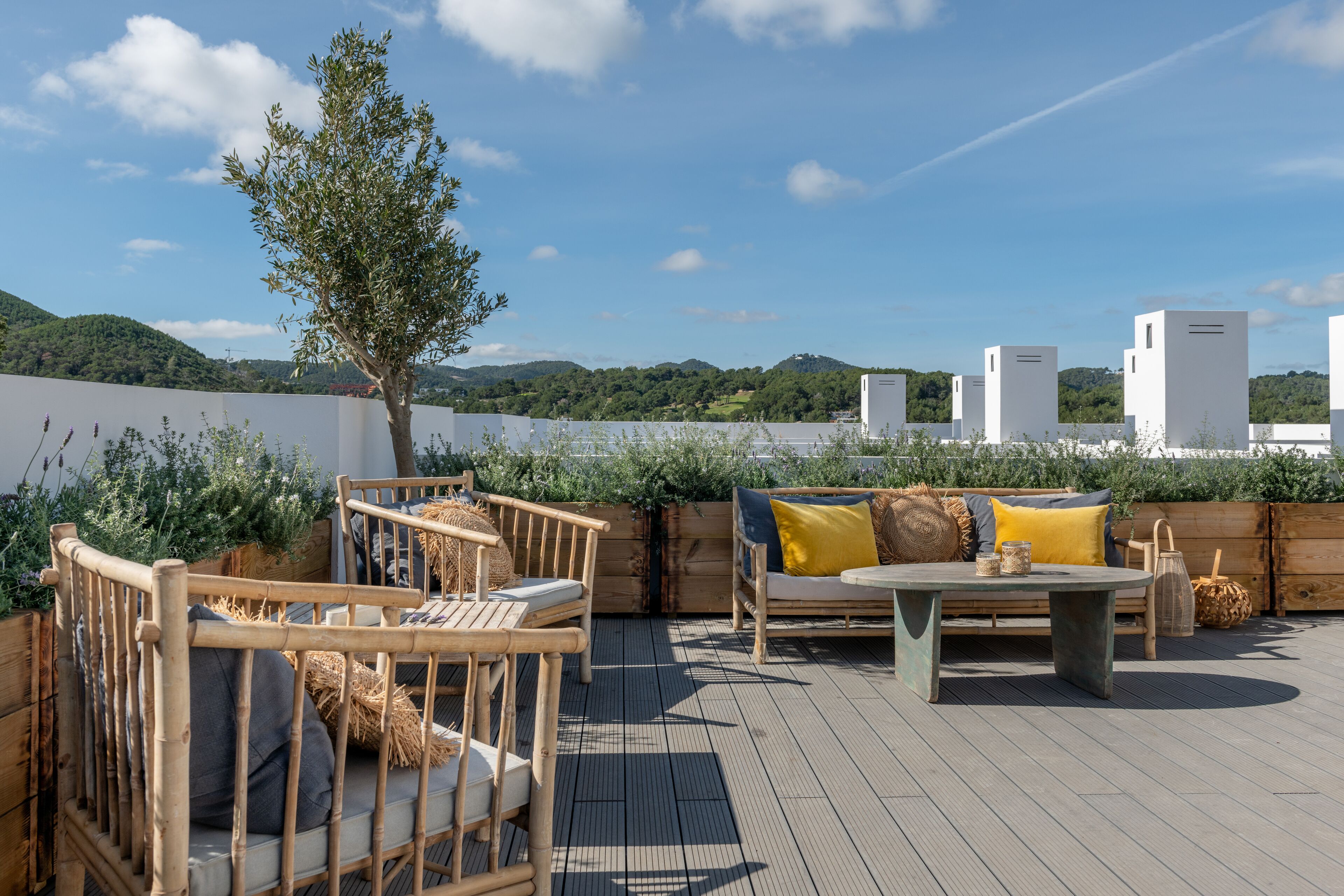 Una terraza al aire libre con mobiliario de madera, una mesa y vistas a colinas verdes y edificios blancos al fondo.