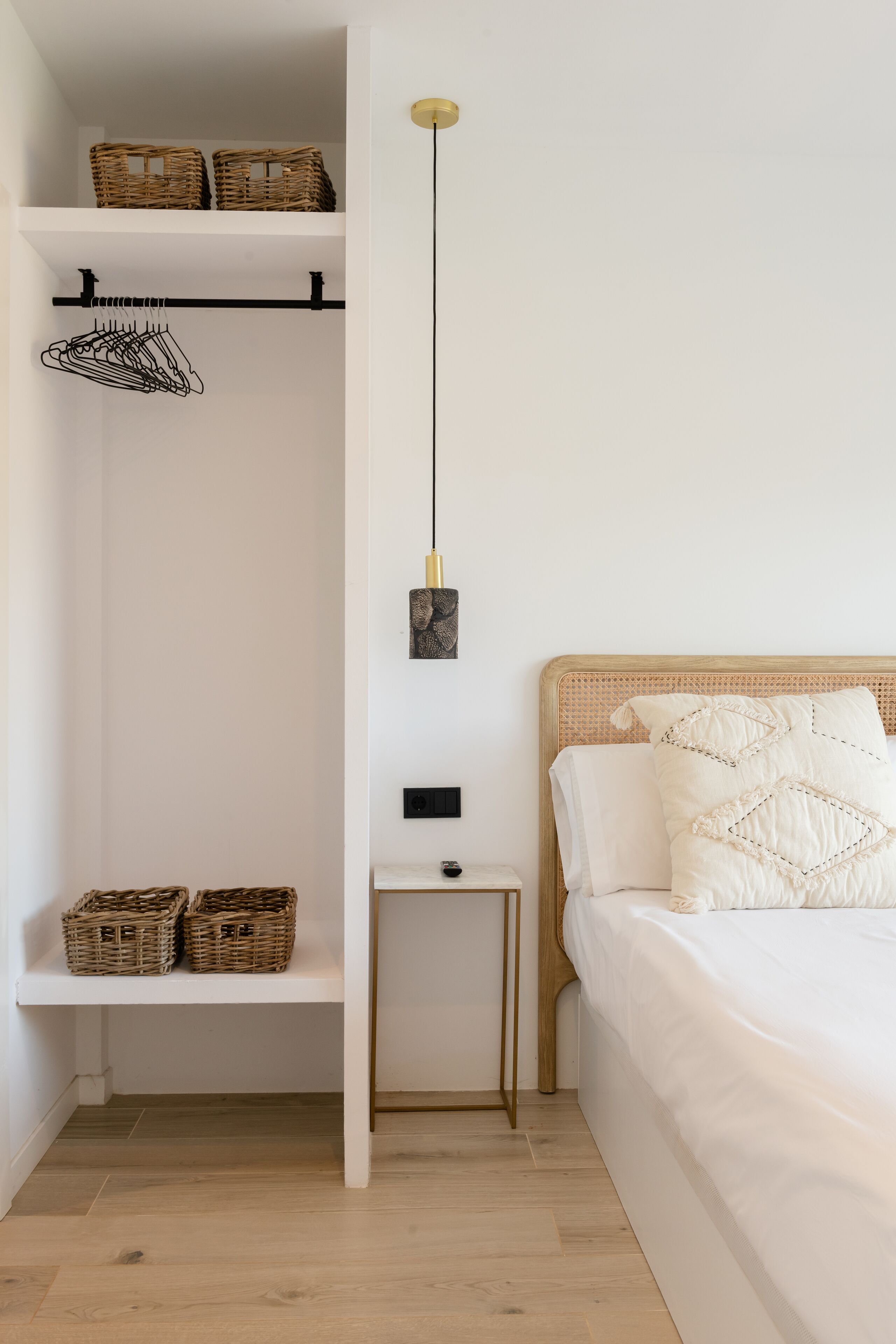 Un dormitorio con cama, mesita de noche, lámpara colgante, estante con cestas y armario con perchero.