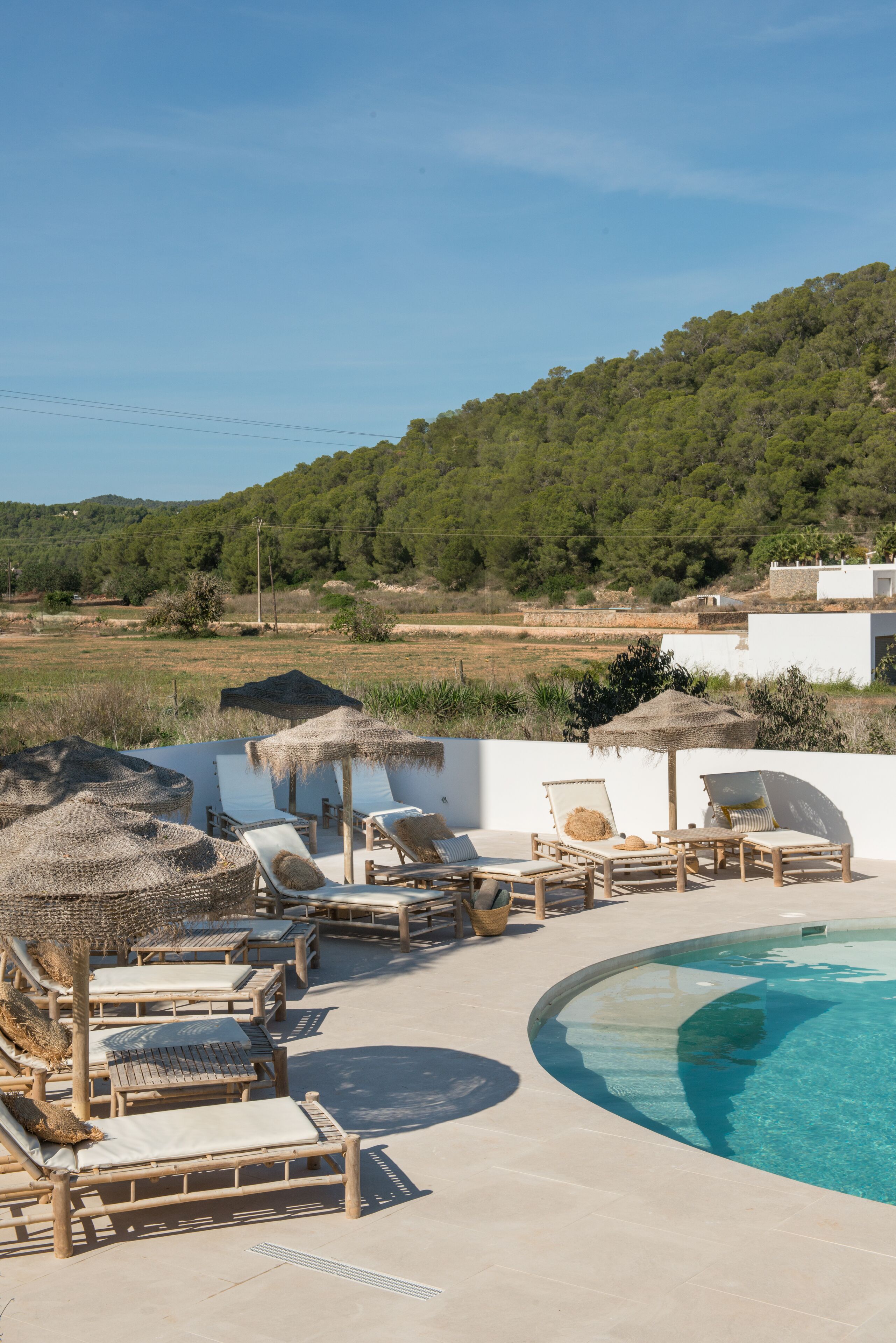 Zona de piscina con sillones y sombrillas, rodeada por un edificio blanco y un paisaje montañoso.