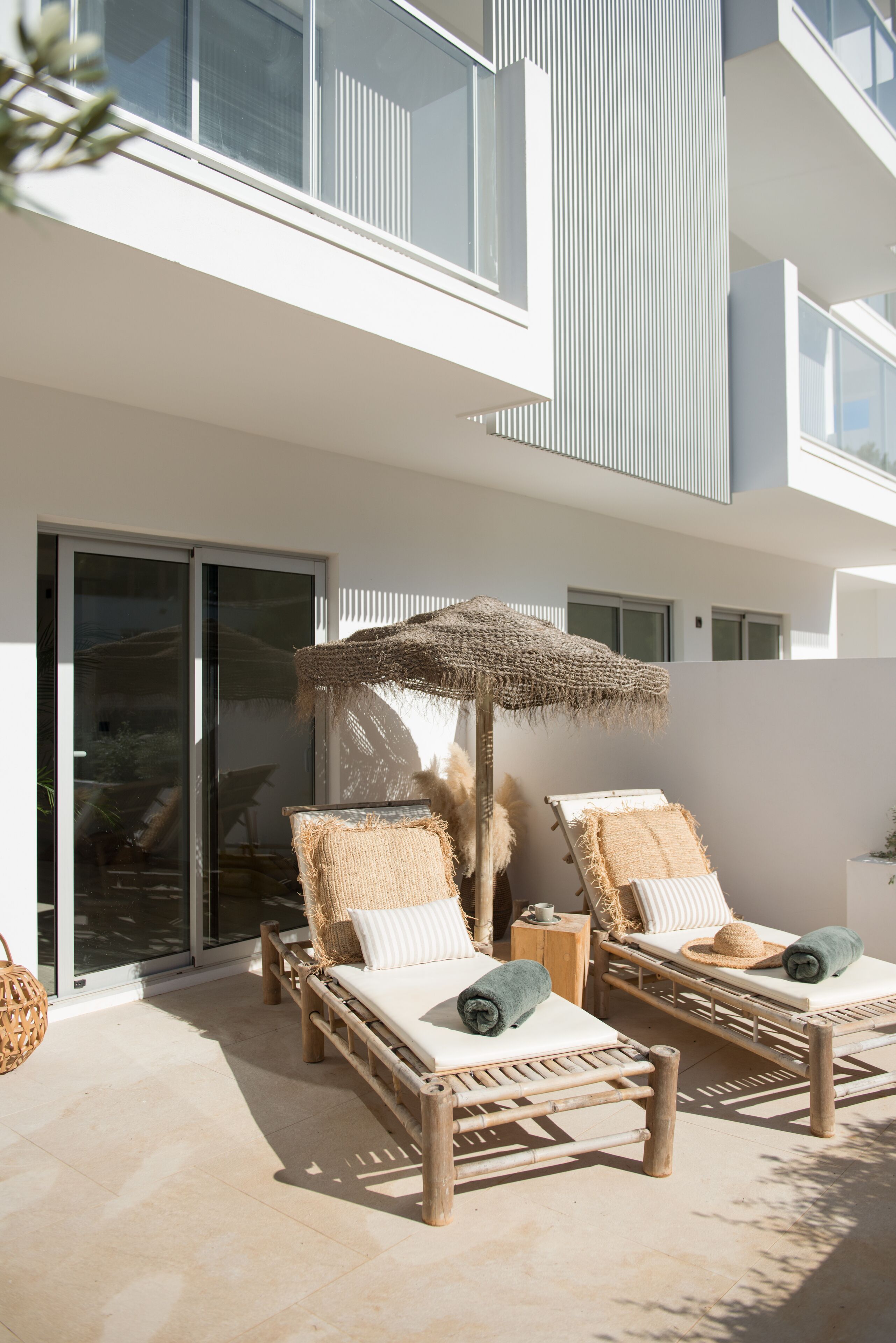 Ein moderner Außenbereich mit einer Lounge-Ecke, zwei Holzliegen, einem gewebten Sonnenschirm und einem Glastür-Eingang.