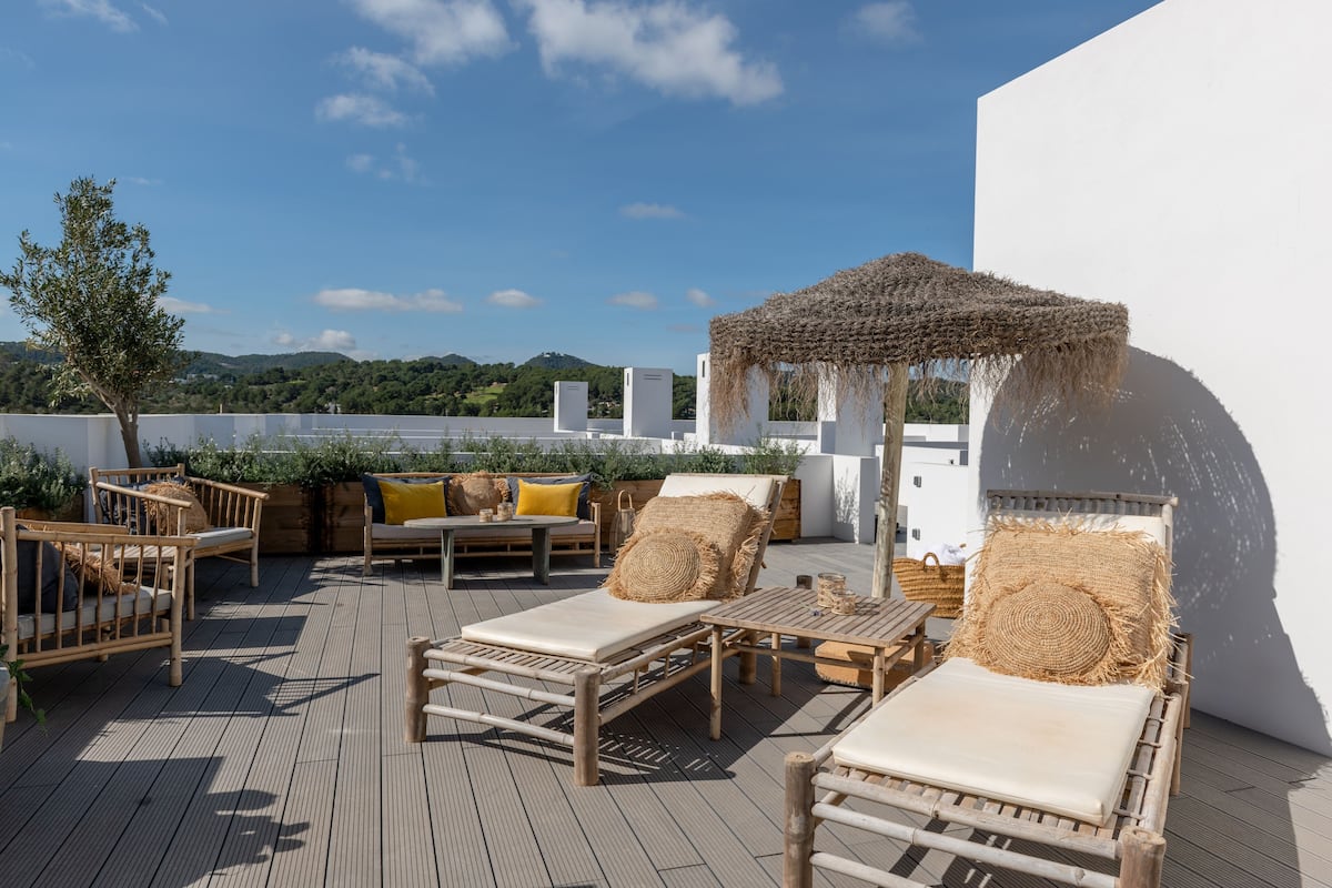 A rooftop terrace with wooden furniture, a thatched umbrella, and a view of greenery and hills.