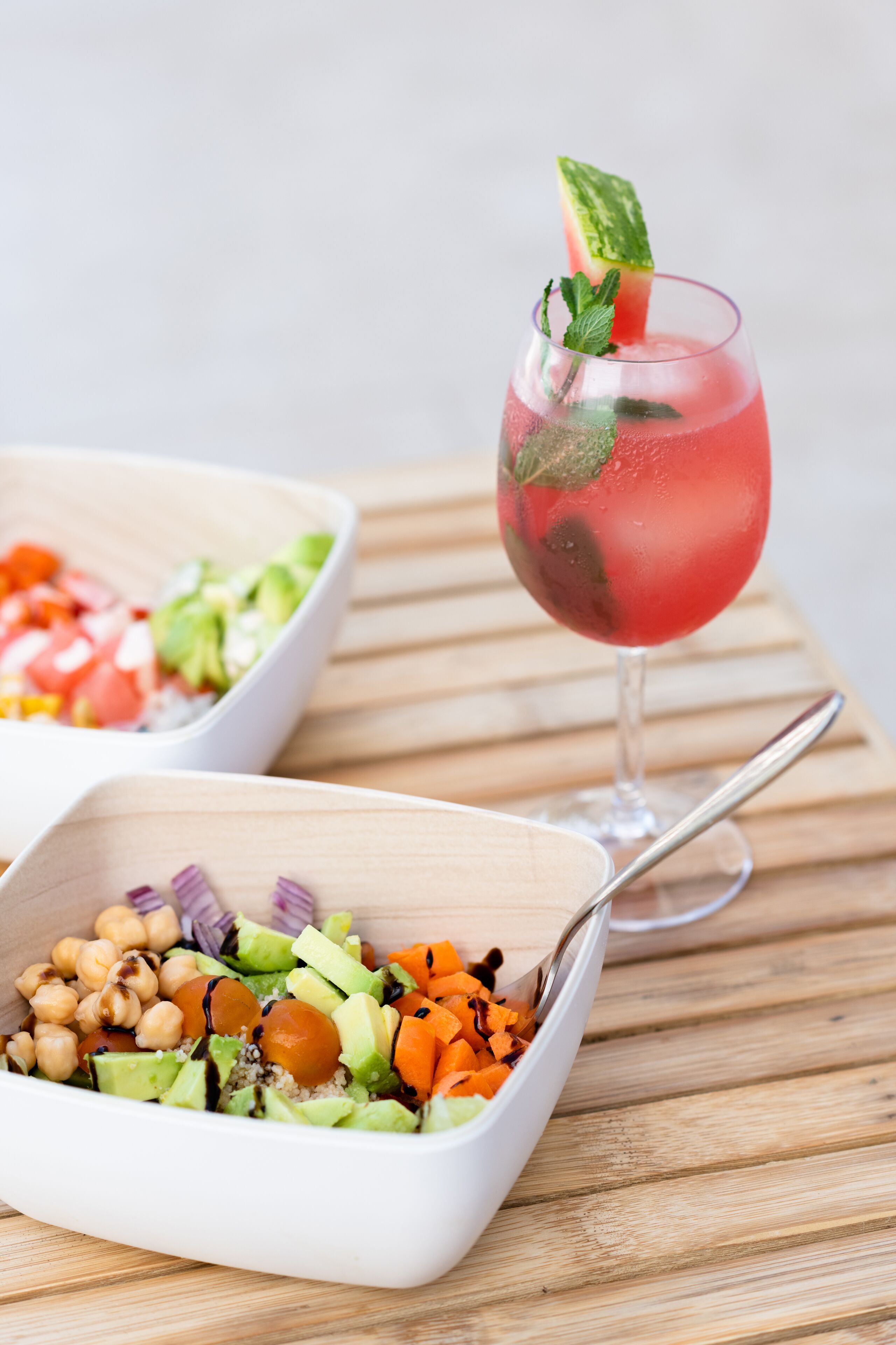 A wooden table with two white bowls containing salads and a glass of red drink with a mint garnish.