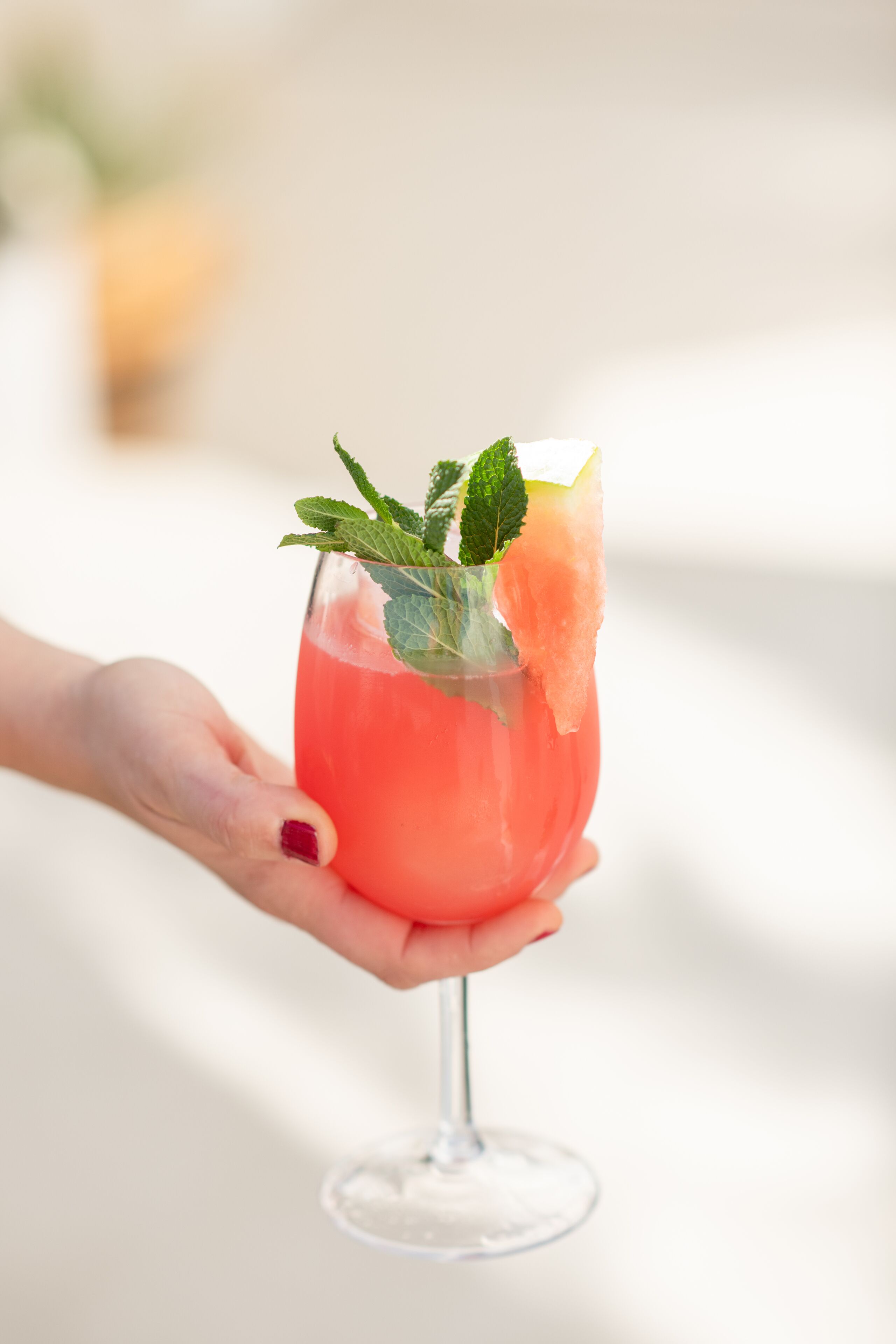A hand holding a glass with a pink beverage, garnished with mint leaves and a slice of fruit.