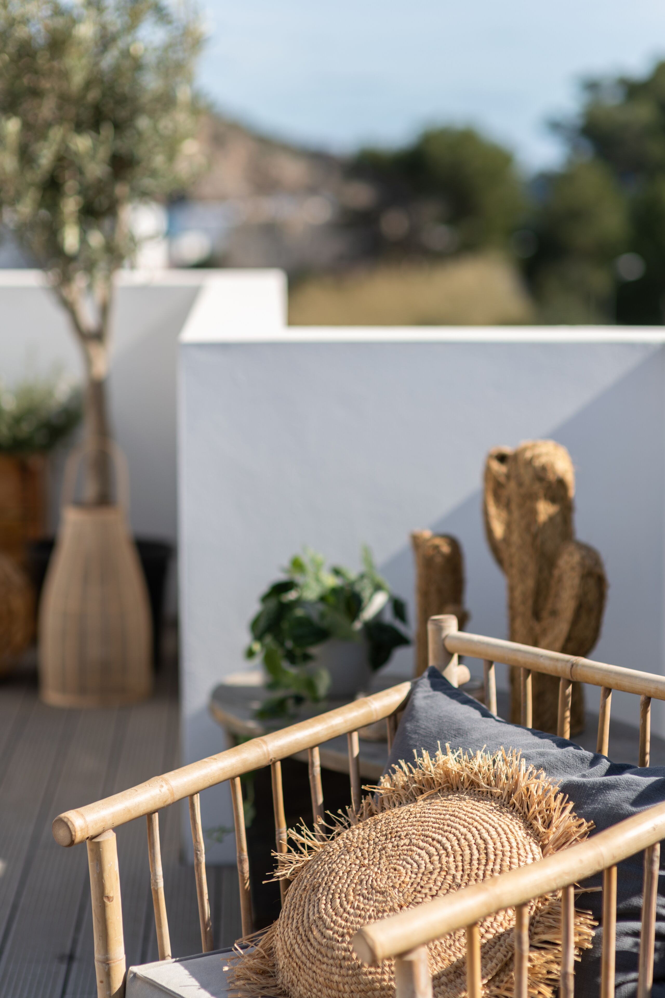 Una silla de mimbre con un cojín tejido y una almohada oscura, sobre una terraza de madera con plantas en macetas y una escultura rústica al fondo.
