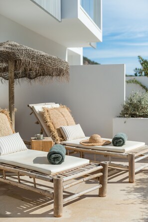 A sunlit outdoor seating area with two lounge chairs, a woven umbrella, and a small table with a cup. - The Olive Boutique Suites and Spa (Santa Eulalia del Rio)