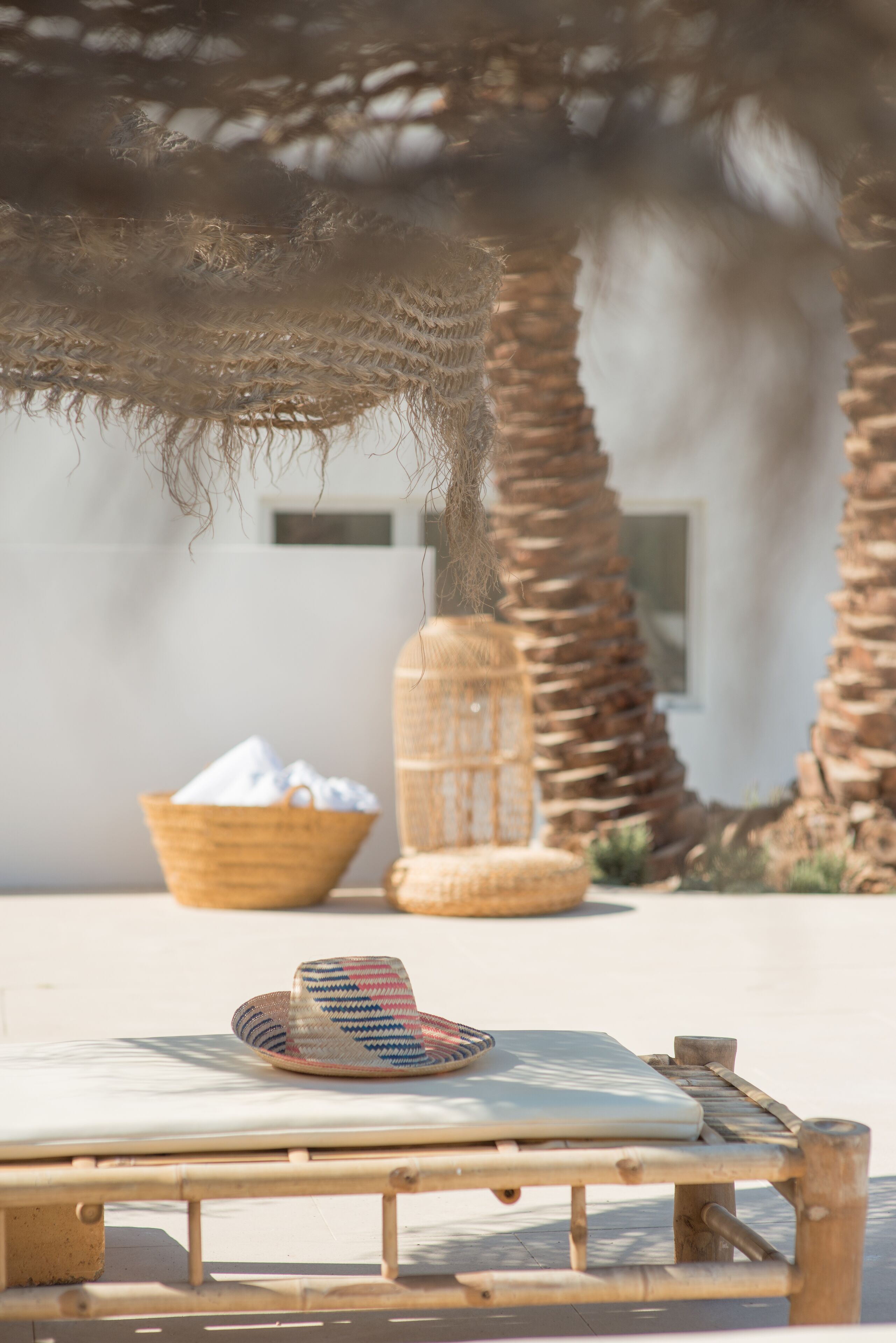 Una tumbona con un sombrero tejido y una cesta de toallas blancas, bajo una palmera en un entorno exterior soleado.