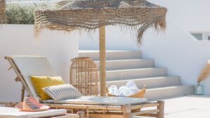 A sun lounger with a woven umbrella, a wicker basket with towels, and a view of a white building with steps. - The Olive Boutique Suites and Spa (Santa Eulalia del Rio)