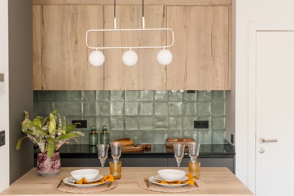 A modern kitchen with wooden cabinets, a green tiled backsplash, and a dining table set with plates, glasses, and a plant.