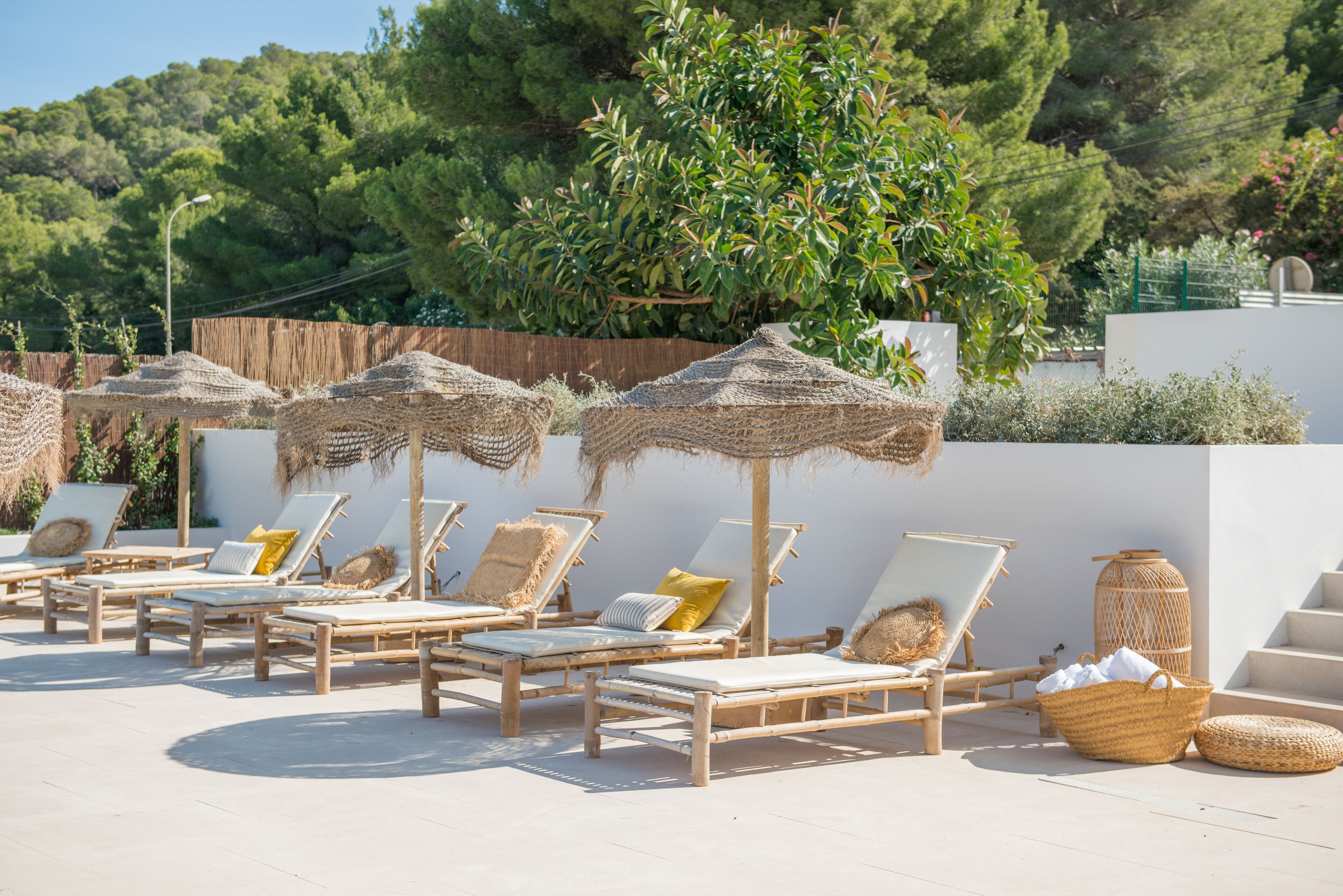 A sunlit outdoor lounge area with wooden lounge chairs and straw umbrellas, surrounded by greenery and a white wall.