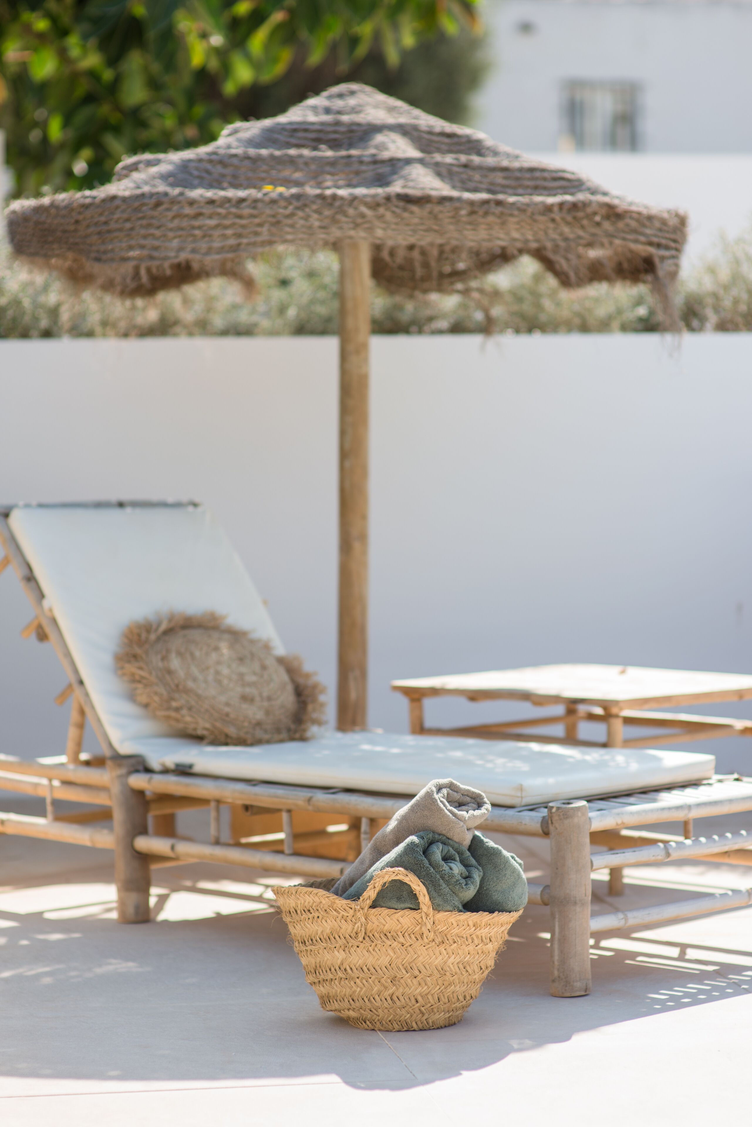 A beachside lounge area with a straw umbrella, a lounge chair with a cushion, and a woven basket with towels.
