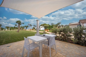 An outdoor patio with a table and chairs, a stone floor, and a view of a garden with trees and a building in the background.