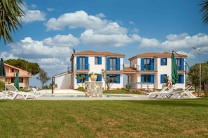 A two-story building with a pool, white walls, blue shutters, and a stone fountain.
