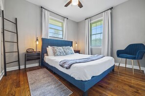 A bedroom with a large bed, a nightstand, a chair, a ladder, and a window with curtains.