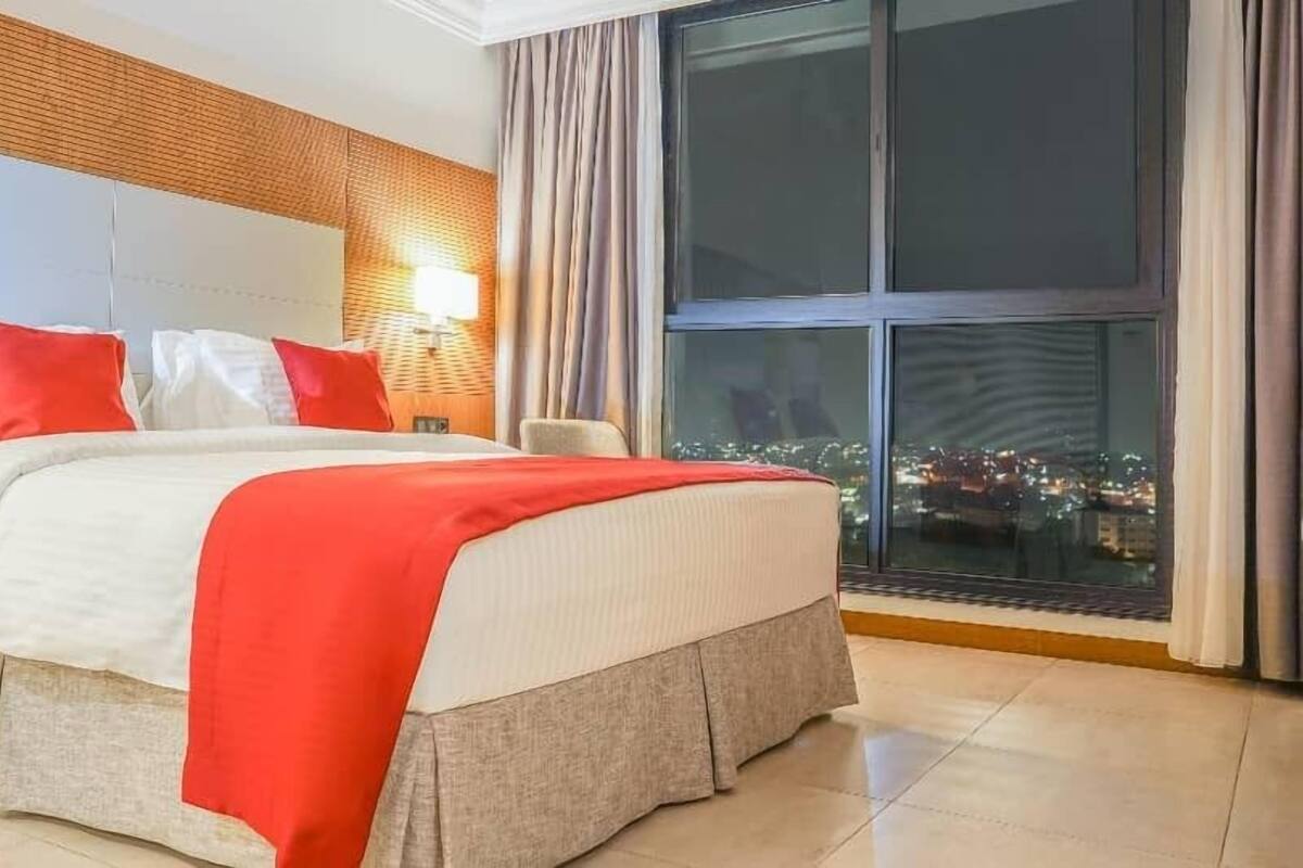 A hotel room with a large bed, a city view through the window, and a modern design with a wooden accent wall.