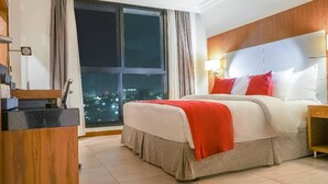 Habitación de hotel con una cama grande, una mesita de noche, un escritorio con lámpara y vista a la ciudad de noche.
