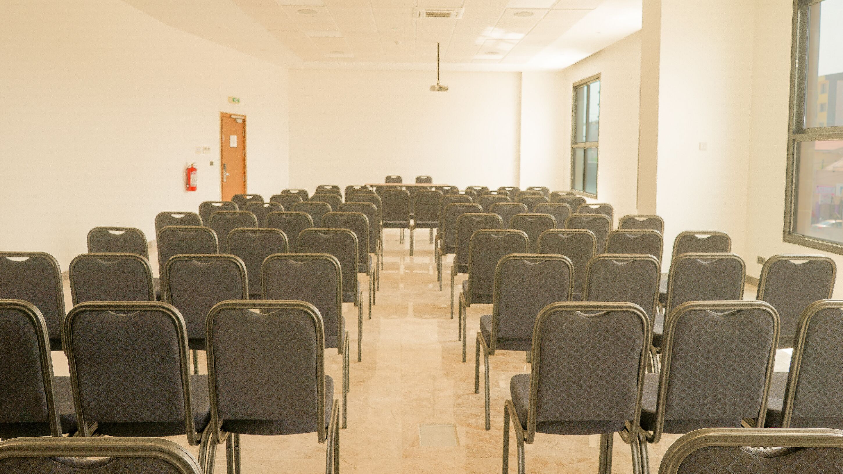 A conference room with rows of chairs facing a long table, a fire extinguisher on the wall, and a window on the right side.
