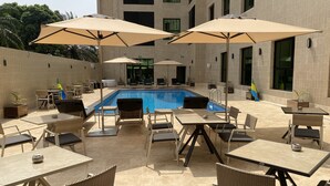 Zona junto a la piscina con mesas, sillas, sombrillas y una piscina.