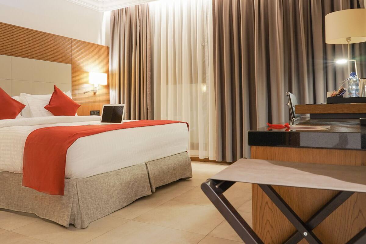 A hotel room with a large bed, a desk with a lamp, and a view of the outside through curtains.