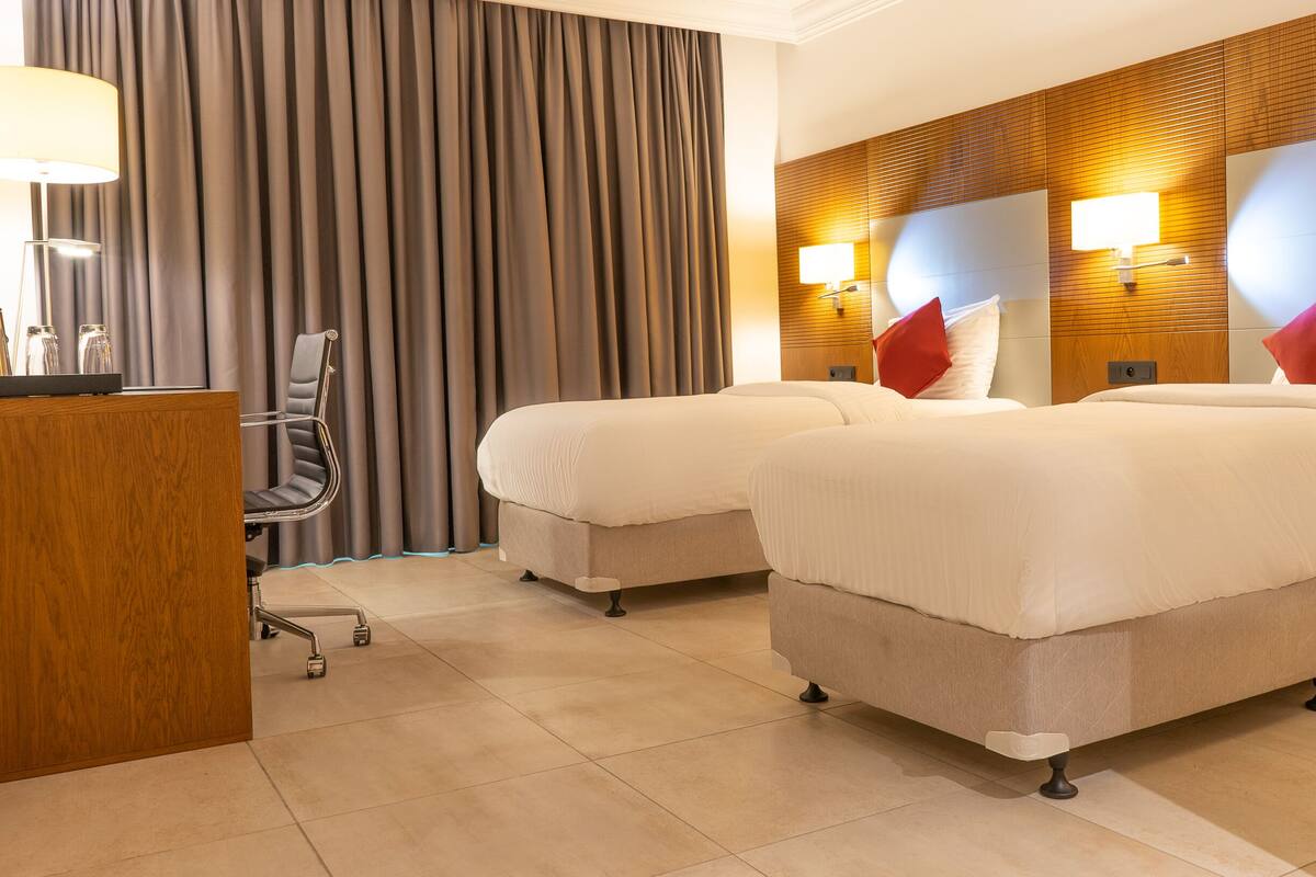 A hotel room with two beds, a desk, a chair, and a large window with curtains.