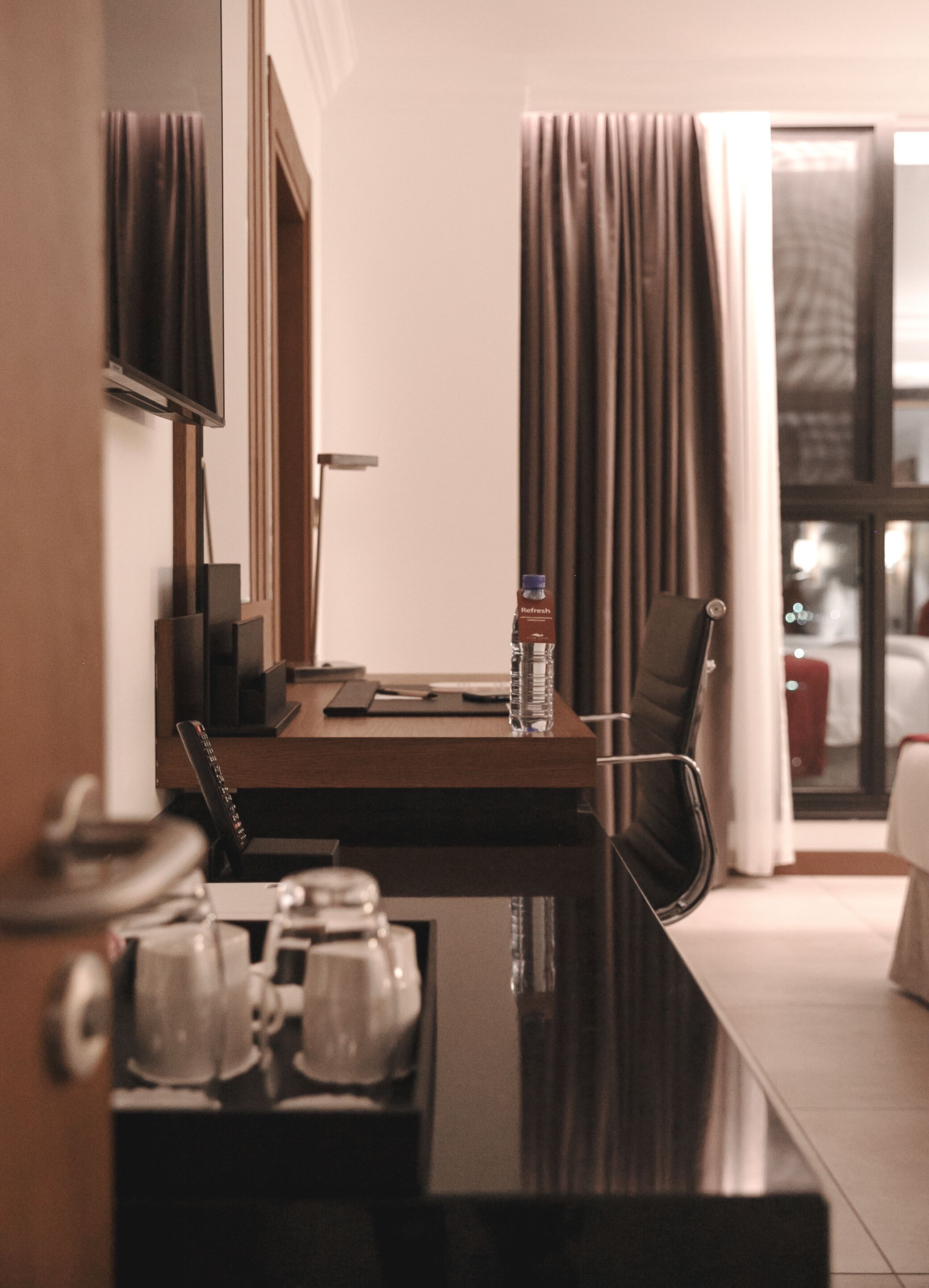 A hotel room with a desk, a chair, a TV, a bottle of water, and a window with curtains.